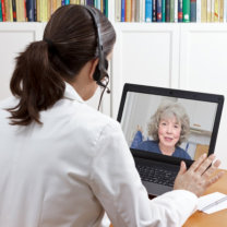 Female doctor of geriatrics in her surgery office with headset in front of her laptop talking via video call with an old patient